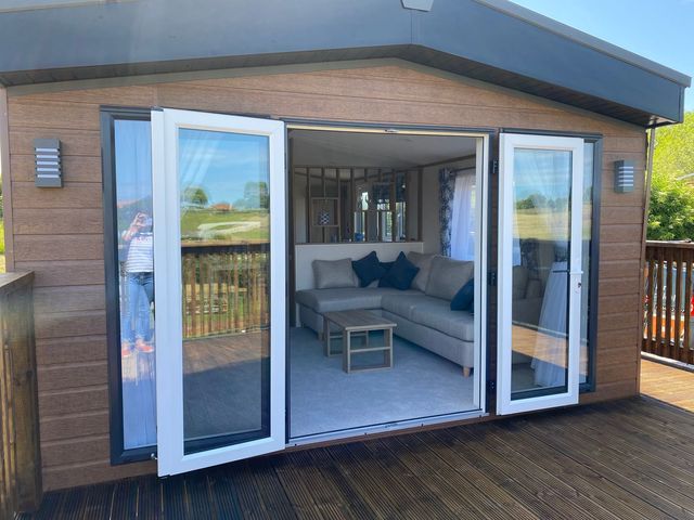 A patio with open glass doors leading to a living room with a grey sofa and wooden coffee table at Maple Lodge in Whitby