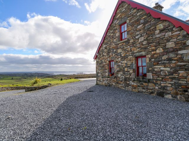 A stone house with red framed windows beside a gravel driveway overlooking green fields at Kates Cottage near Clonbur County Galway