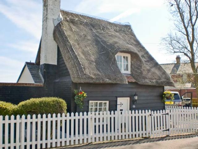 A small thatched roof cottage with a white picket fence and hanging flower baskets at Little Thatch in Walton-On-The-Naze