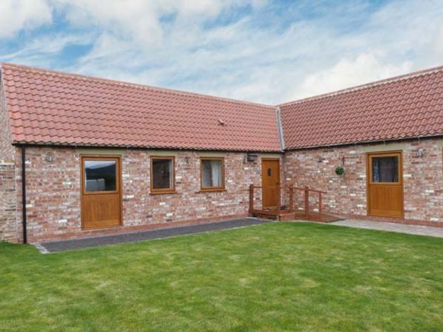 A grass yard in front of a brick building with three wooden doors and two windows at Cooper Cottage in Potto near Stokesley