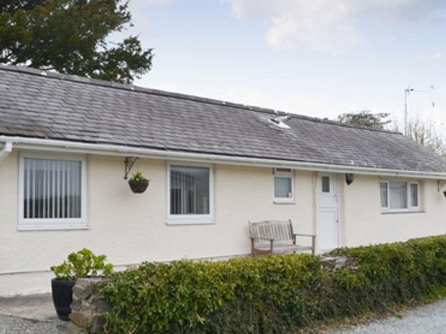 A single-story house with white walls and a gray roof with a bench outside and trimmed bushes in front at Plas Cottage in Llanfaelog