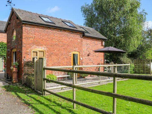 A small red brick building with wooden framed windows and skylights a wooden fence and gate grass and trees at Chequer Stable in Sandbach