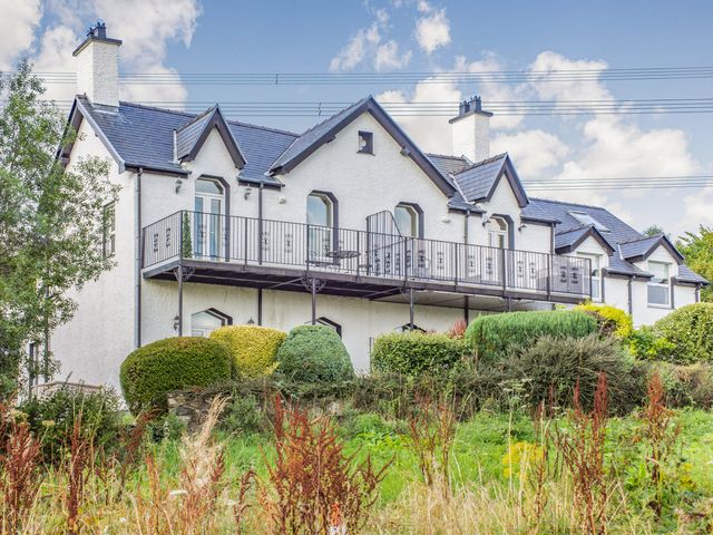 A white two-story house with a black roof and a balcony surrounded by bushes and grass at Menai View in Llanfairpwllgwyngyll near Beaumaris