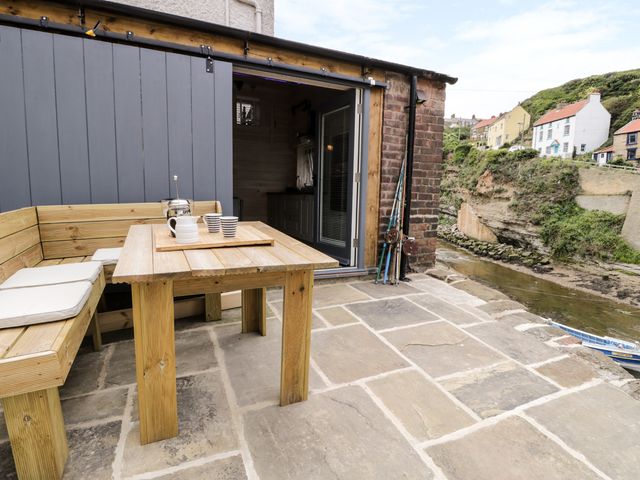 An outdoor patio with wooden table and bench seating next to a sliding door overlooking a river and houses at Old Joiners Shop Staithes