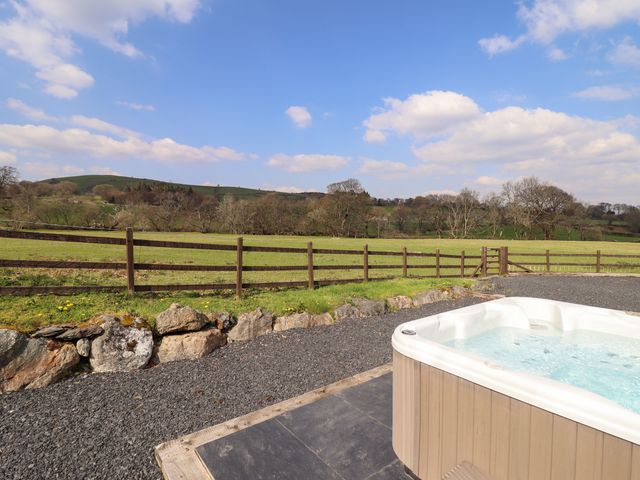 A hot tub on a patio with a gravel area and a fenced grassy field with trees and hills at Beudy Bach Barn in Llanuwchllyn near Bala