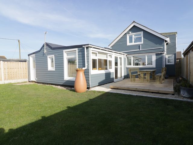 A backyard with a lawn wooden deck table and benches and a blue house at Home to Roost in Eccles-On-Sea