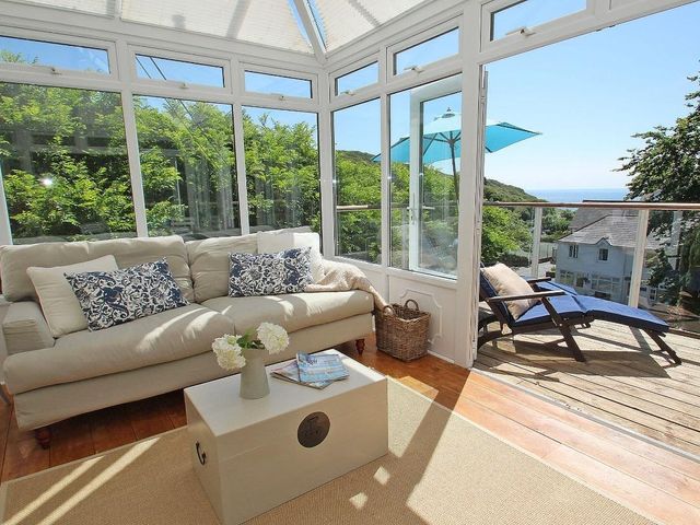 A sunroom with a beige sofa a small table with flowers and magazines and a deck with lounge chairs and an umbrella at Porthcurno Bay View in Porthcurno