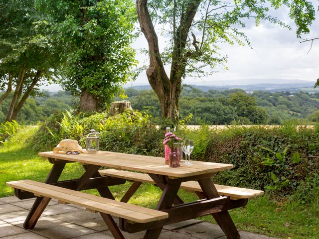 A wooden picnic table with flowers glasses and a hat in a garden with trees and a countryside view at Dartmoor View in Gunnislake