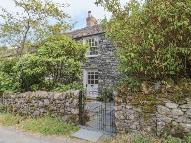 A stone cottage with a white door and window behind a stone wall and iron gate surrounded by bushes at Stair Cottage in Stair nr Keswick Cumbria