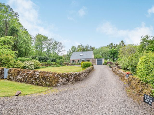 A house with a gravel driveway and stone walls surrounded by trees and greenery at Falla Farmhouse in Jedburgh
