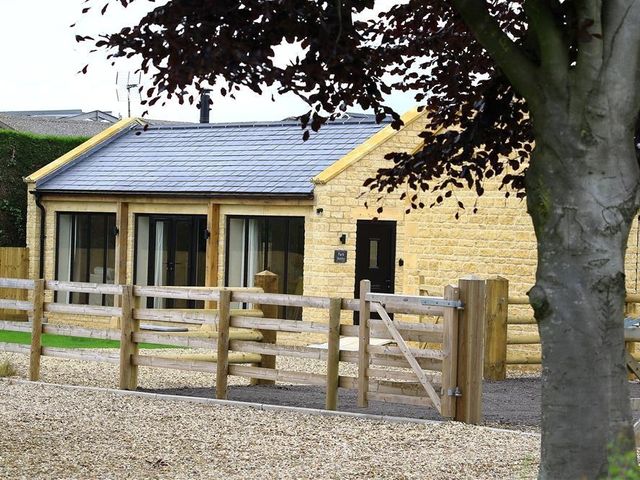 A stone building with a slate roof behind a wooden fence and a tree at Park Bothy in Wyck Rissington