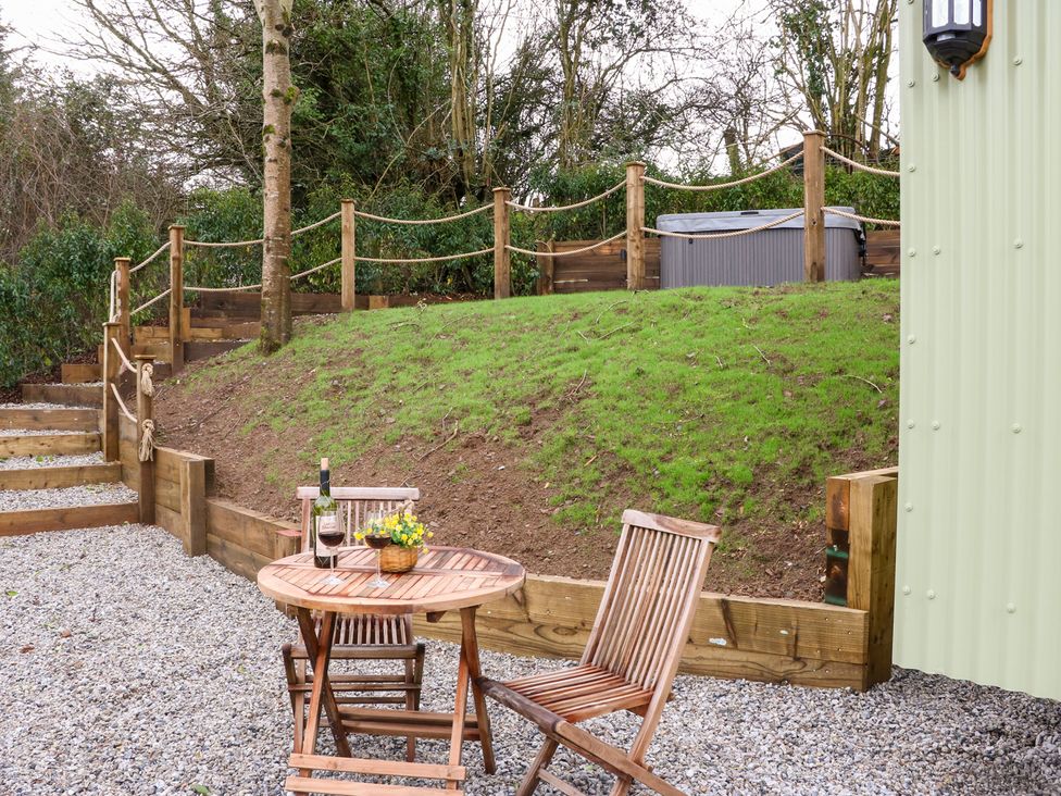 An outdoor patio area with a wooden table and two chairs a bottle of wine and two glasses on the table grass hill with rope railing and steps leading up to a hot tub at Little Silver Leaf in High Bickington