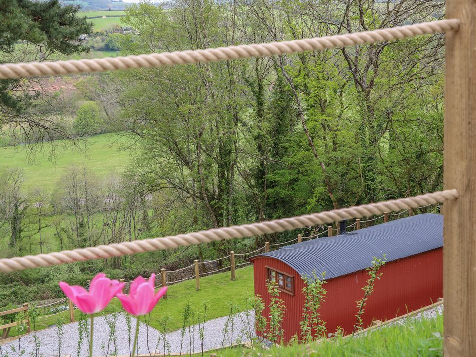 A red corrugated building with a curved roof near a green landscape with trees and pink flowers in the foreground at Little Silver Owl in High Bickington