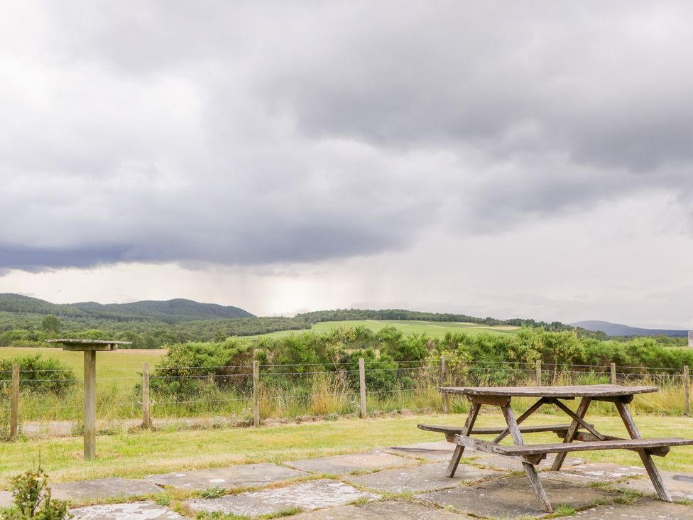 A wooden picnic table on a stone patio with a fenced grassy field and hedges under a cloudy sky