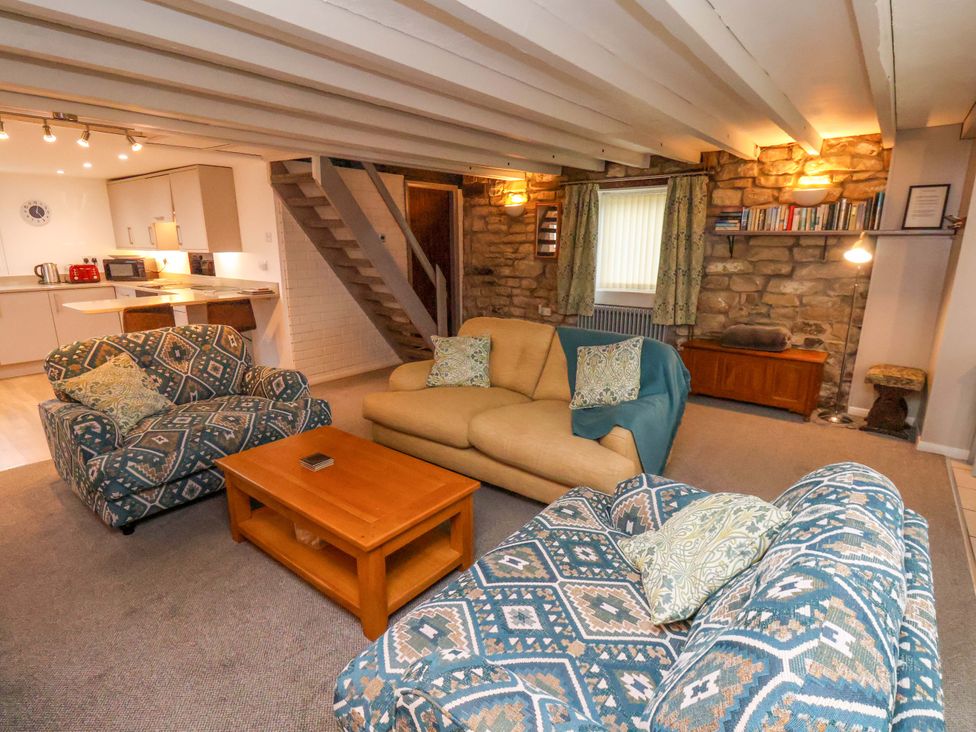 A living room with patterned armchairs a beige sofa and a wooden coffee table next to a kitchen area with stairs and a bookshelf on a stone wall at Orchard Cottage in Robin Hoods Bay