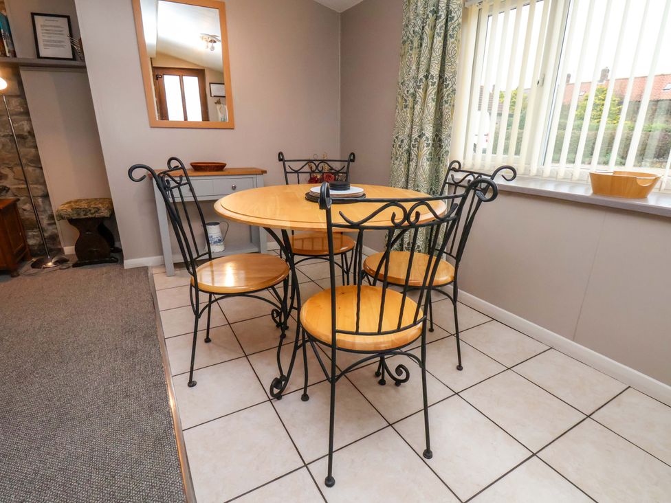 A dining area with a round wooden table and four metal chairs next to a window with vertical blinds and patterned curtains at Orchard Cottage in Robin Hoods Bay