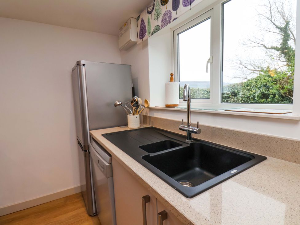 A kitchen countertop with a black sink a silver refrigerator and kitchen utensils in a holder by a window at Orchard Cottage in Robin Hoods Bay