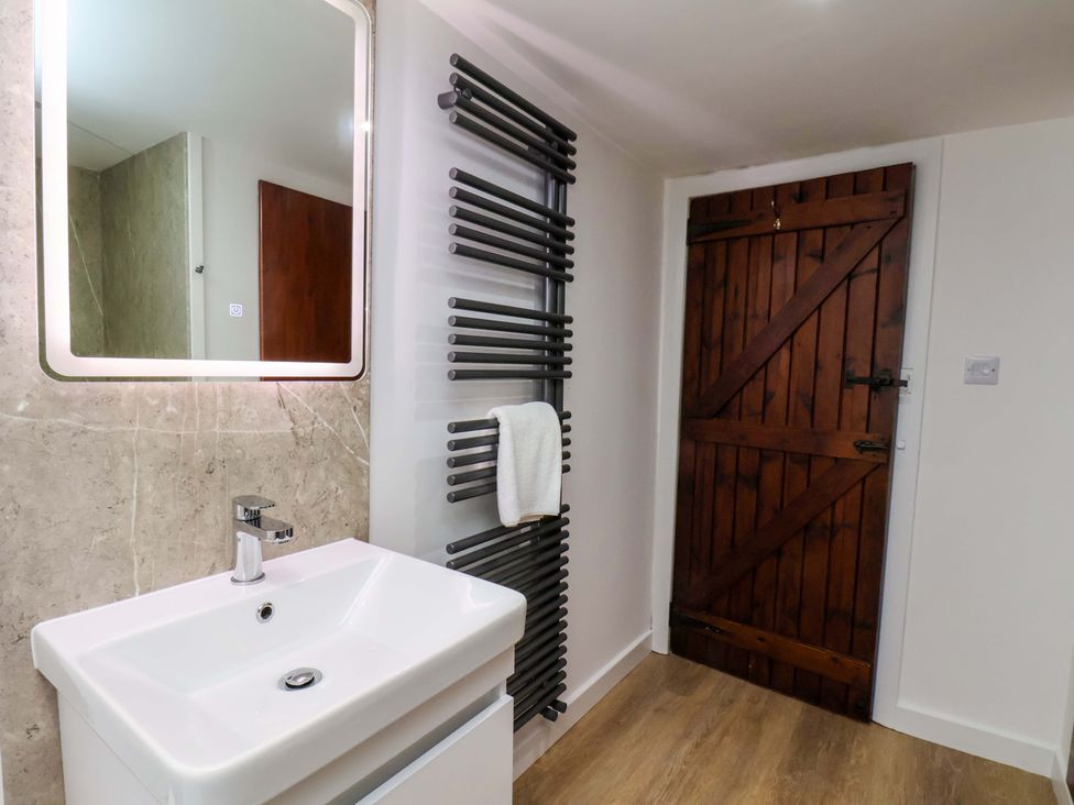 A bathroom with a white sink and modern faucet a backlit mirror and a black heated towel rail with a white towel and a wooden door at Orchard Cottage in Robin Hoods Bay