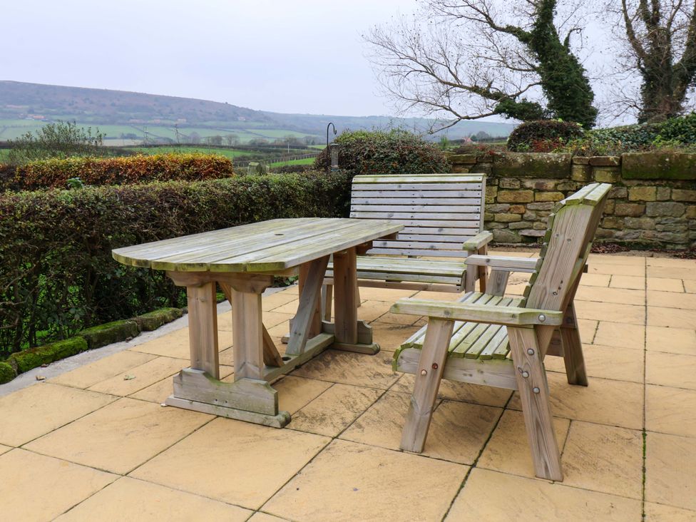 A wooden outdoor table with two wooden benches on a tiled patio with hedges and a stone wall in the background