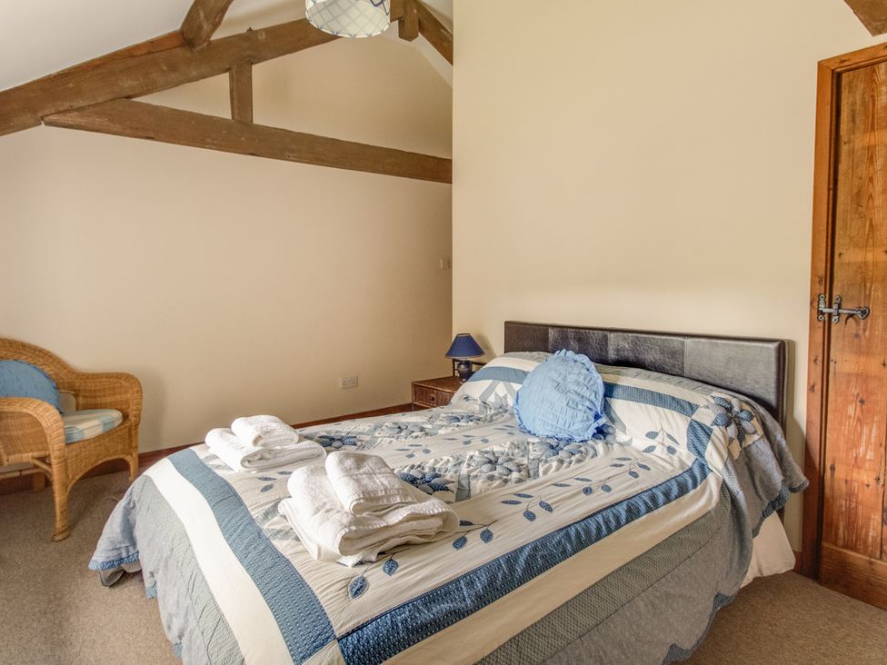 A bedroom with a double bed covered with a blue and white patterned quilt and folded towels on top with a wicker chair and a wooden door at Churn House in Dewlish