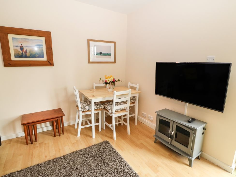 A dining area with a wooden table and four chairs with cushions a television mounted on the wall above a cabinet two framed pictures on the wall and a small set of nesting tables on wooden floor at Plas Marchog Annexe in Rhoscefnhir near Pentraeth