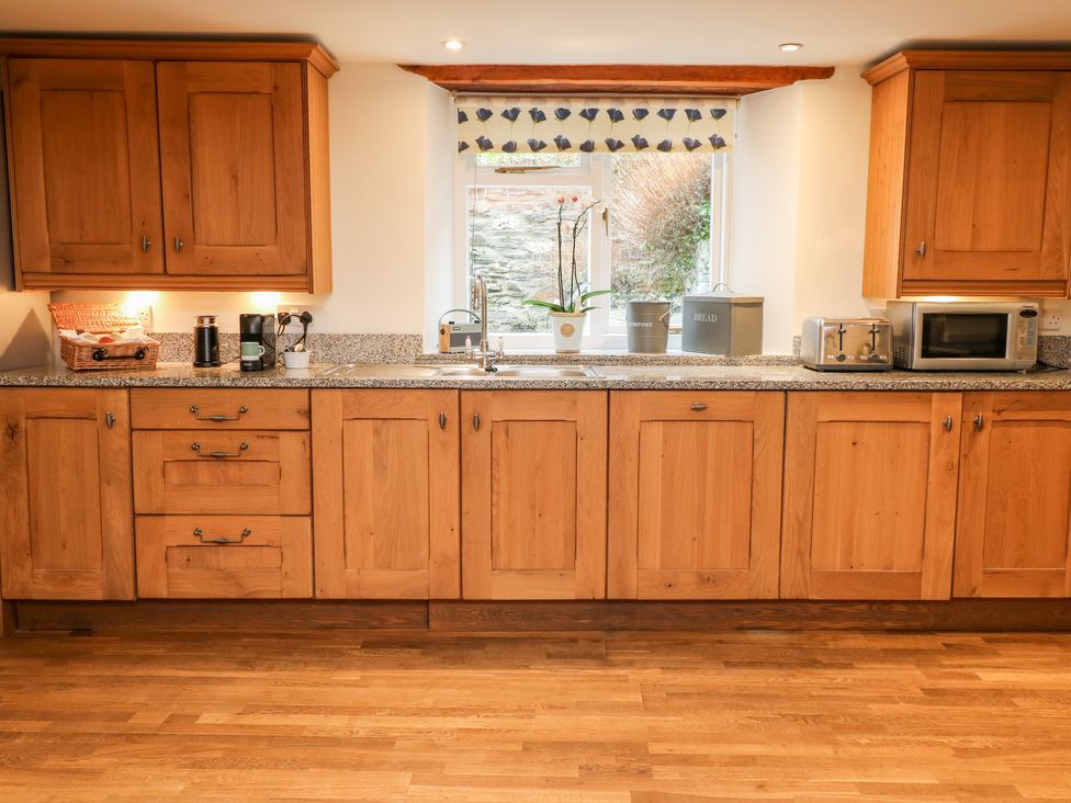 A kitchen with wooden cabinets a window above the sink a toaster microwave and kitchen appliances on the countertop at Court Prior Cottage in Cornworthy