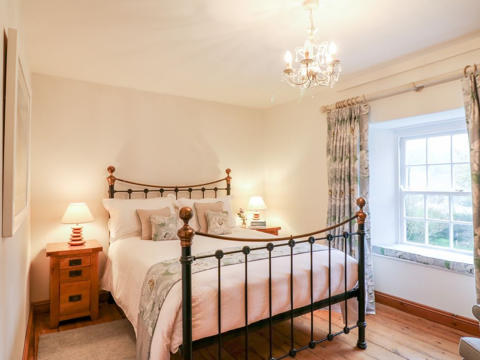 A bedroom with a metal bed frame wooden bedside tables lamps floral curtains and a window at Court Prior Cottage in Cornworthy