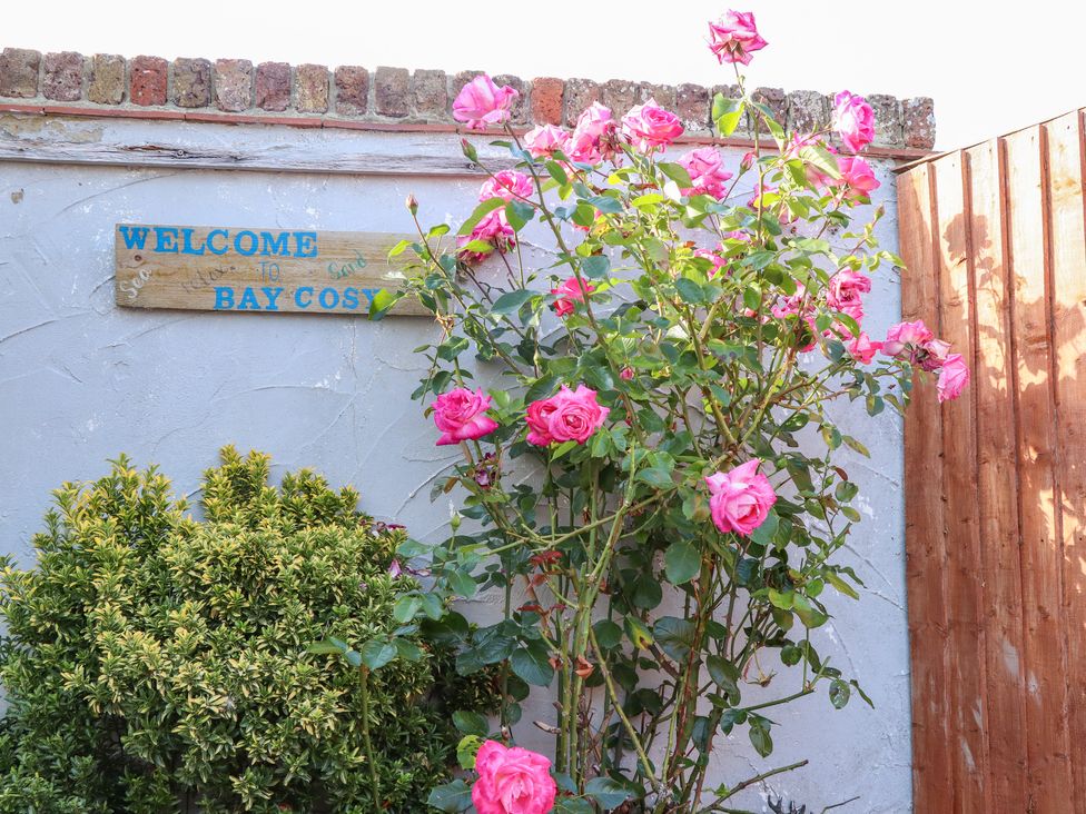 A garden wall with a wooden welcome sign and pink roses at Bay Cosy in Pevensey Bay