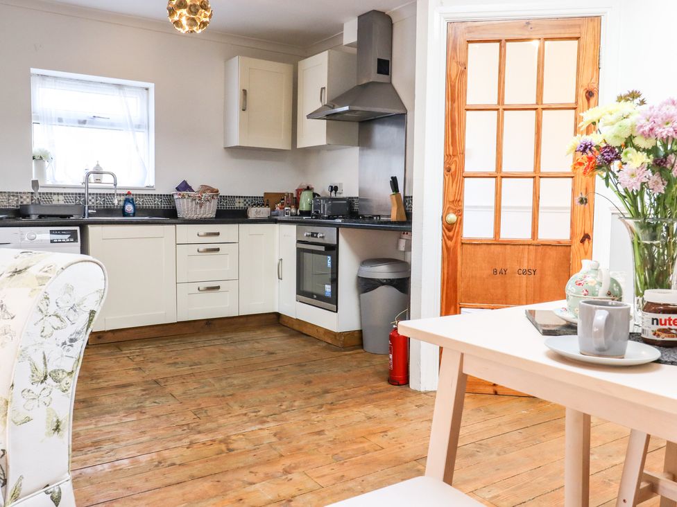 A kitchen with white cabinets wooden floor and a table with flowers at Bay Cosy in Pevensey Bay