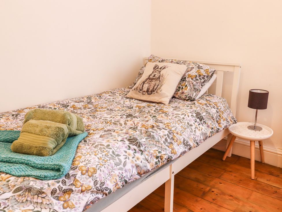 A bedroom with a single bed with floral bedding a pillow with a rabbit design folded towels on the bed and a small side table with a lamp at Bay Cosy in Pevensey Bay
