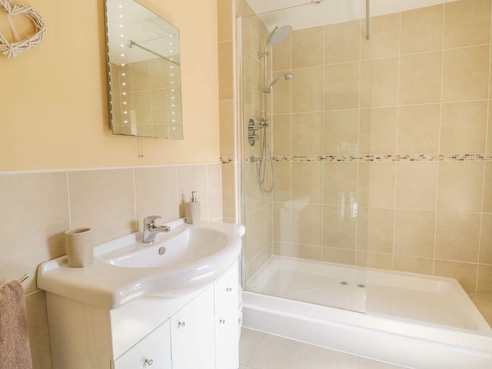 A bathroom with a white sink and cabinet a mirror towel holder and a shower area with tiled walls at Angorfa in Llanbedr