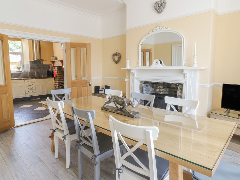 A dining room with a glass top table and chairs near a fireplace and an open door to a kitchen at Angorfa in Llanbedr