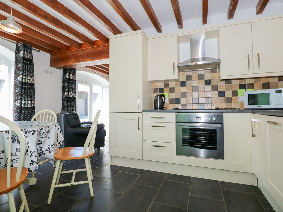 A kitchen with white cabinets a tiled backsplash an oven a microwave kettle and a dining table with chairs and a black armchair at Hafod in Pontllyfni