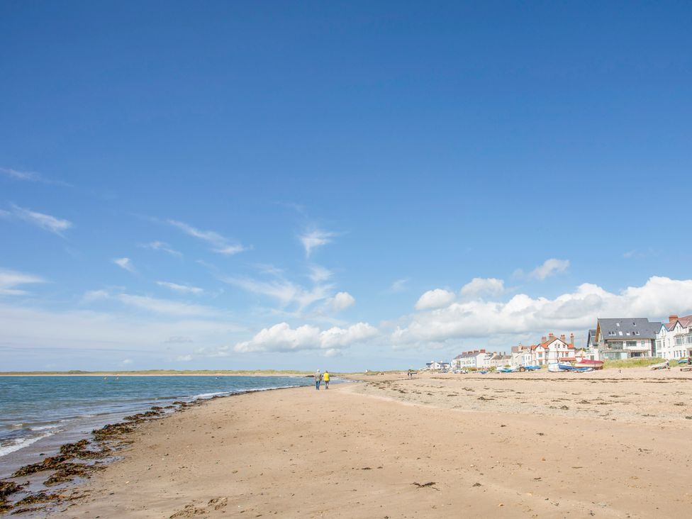 A sandy beach with two people walking near the water and houses along the shore at Windygates in Rhosneigr