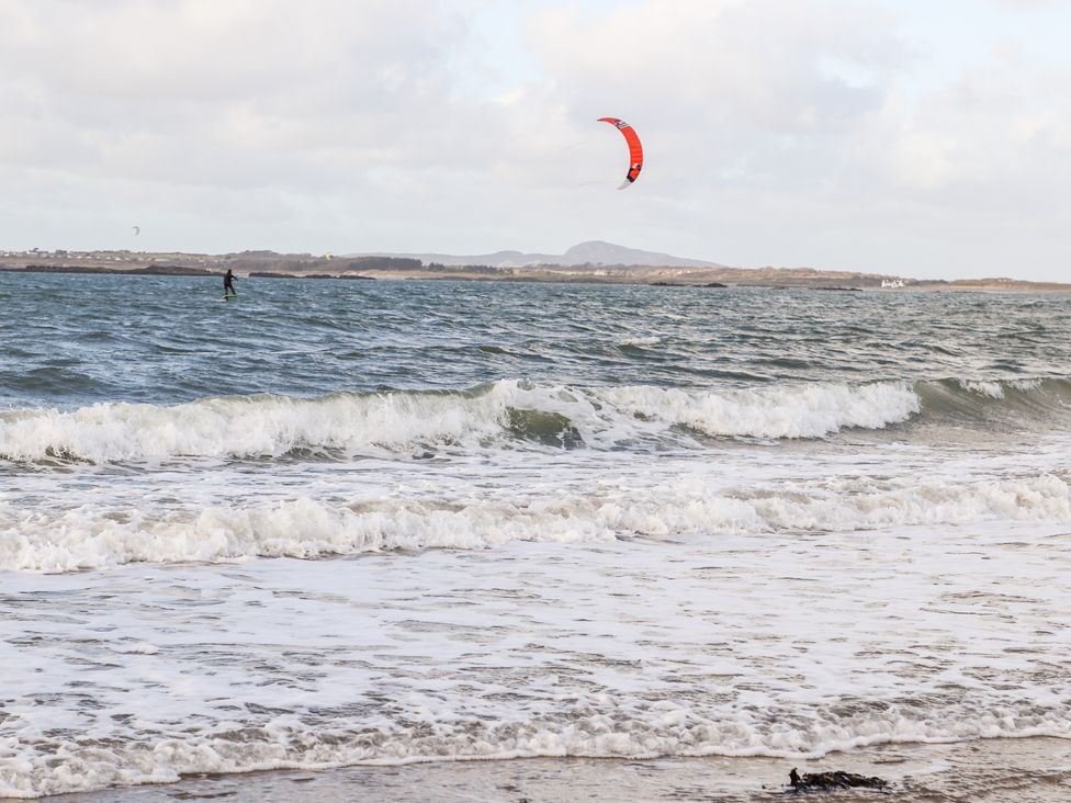 A beach with waves and a person kite surfing on the water with hills in the background at Windygates in Rhosneigr