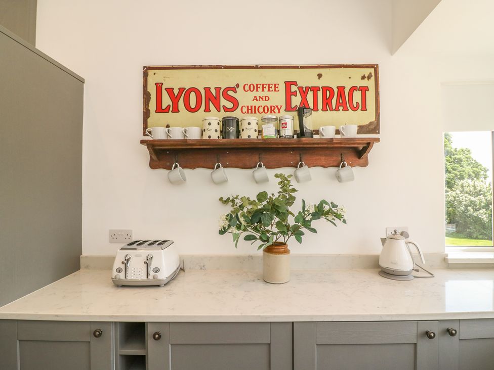 A kitchen countertop with a toaster a kettle a vase with plants and a wooden shelf with cups and jars under a vintage coffee sign at The Coach House in Teignmouth