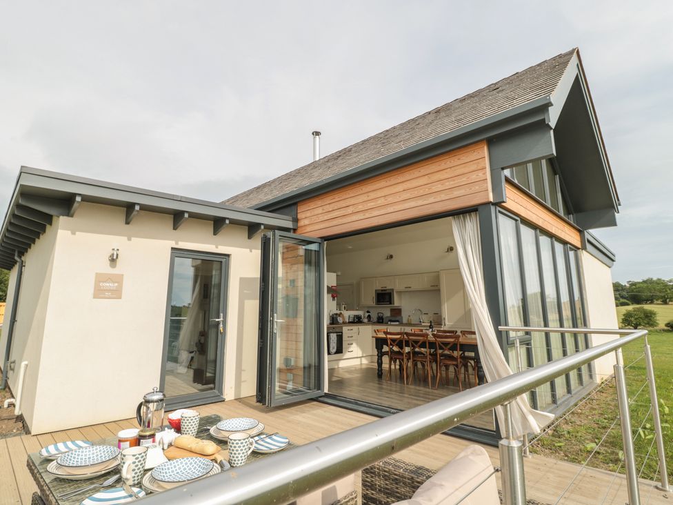An outdoor dining table set with plates and cups on a deck outside a modern house with glass doors open to a kitchen and dining area at Cowslip in Bamburgh