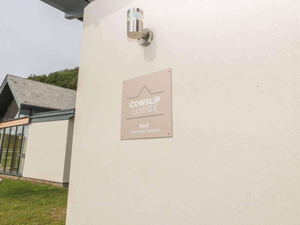 Exterior wall with a sign reading Cowslip Lodge No 6 Brada Lodges Bamburgh and part of adjacent building with grass