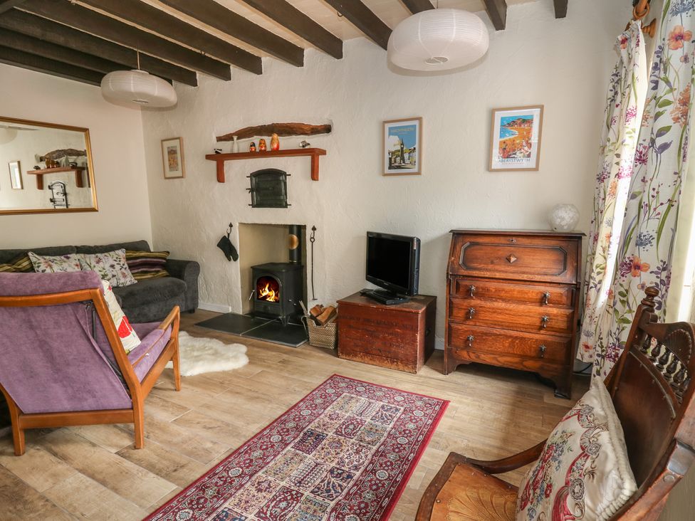 A living room with a wood burning stove a purple armchair wooden furniture and floral curtains at Cysgod Y Coed in Tre'r Ddol