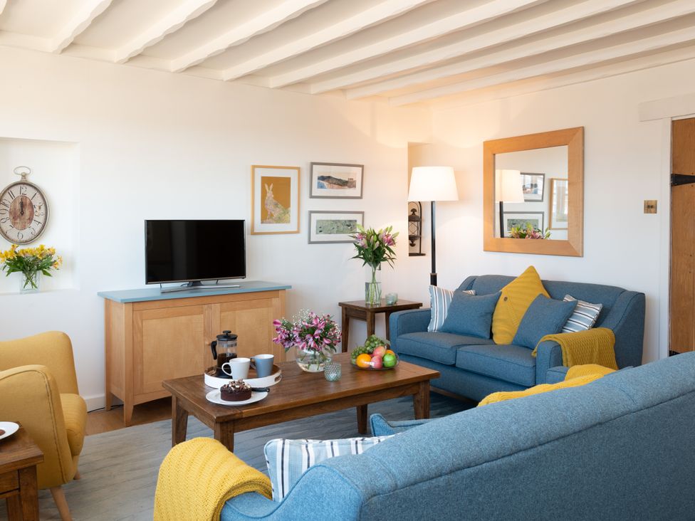 A living room with blue sofas yellow cushions and armchair a wooden coffee table with flowers fruit and drinks and a wooden cabinet with a television at Roxby Cottage in Staithes