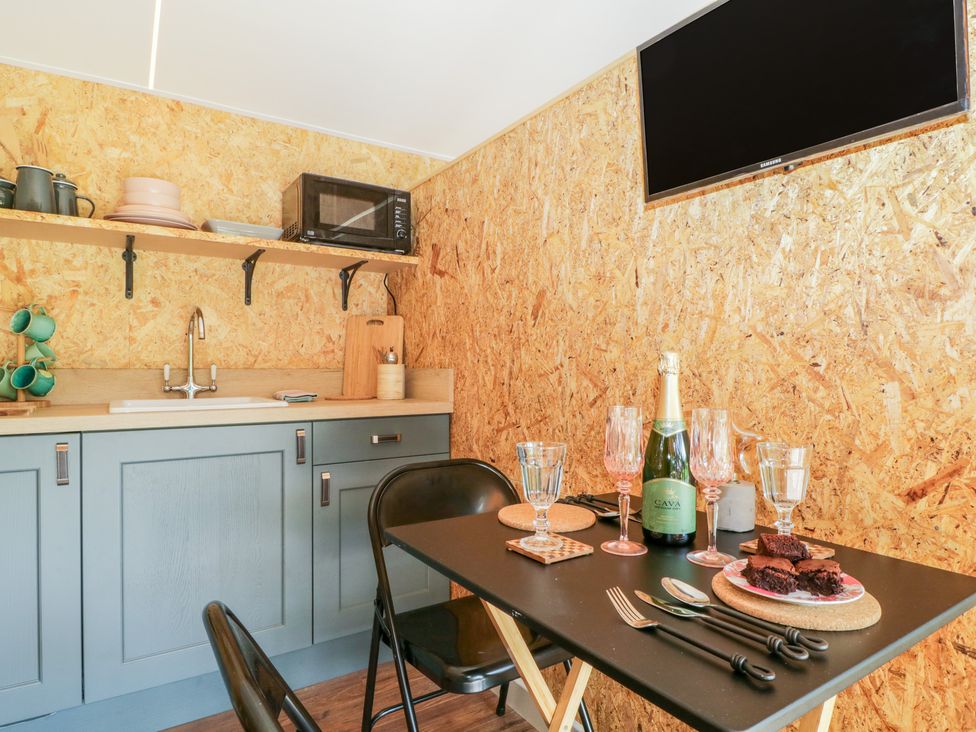 A small kitchen with blue cabinets a microwave on a shelf and a black dining table with glasses a bottle and brownies at The Chard in Longtown