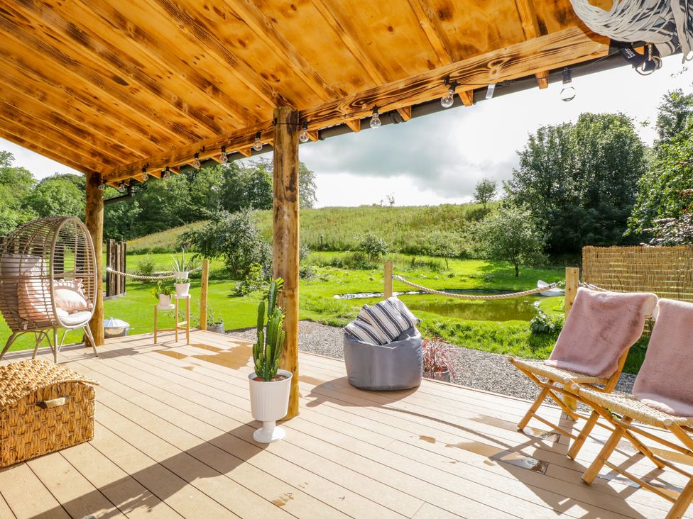 An outdoor covered wooden deck with chairs a hanging chair a bean bag and potted plants overlooking a green lawn and trees at The Chard in Longtown