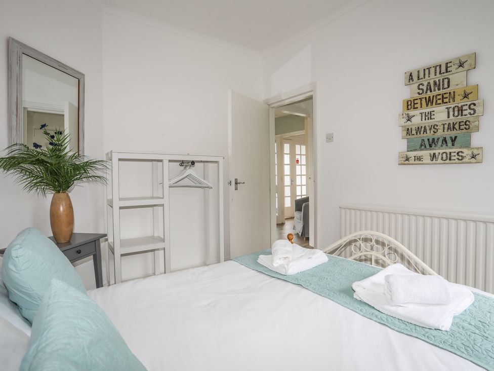 A bedroom with a bed with towels and pillows a small table with a vase and a mirror a white open wardrobe and a wall sign at Casita in Benllech