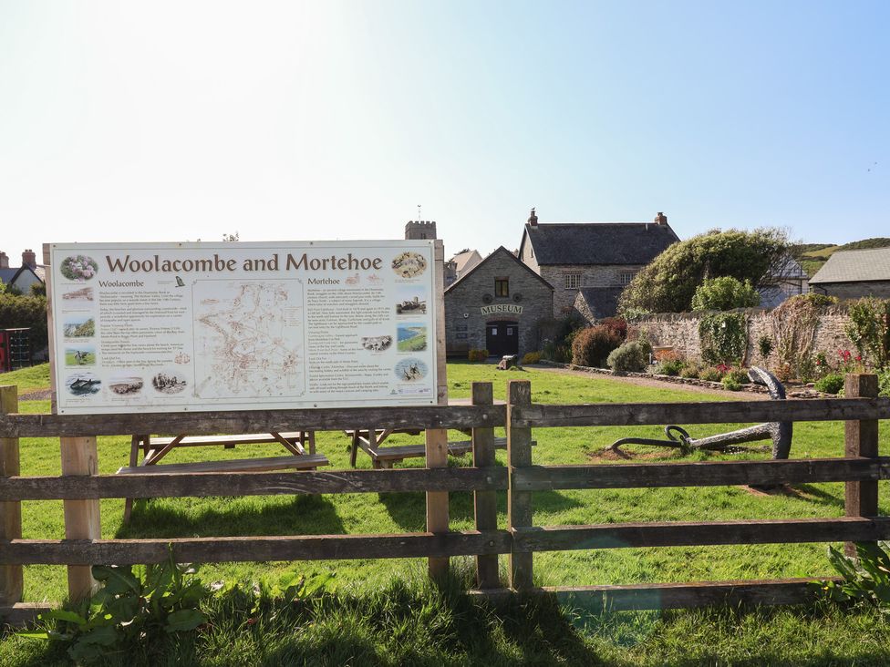An information board about Woolacombe and Mortehoe behind a wooden fence with a museum building and garden in the background