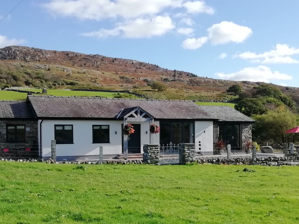 A single story house with a tiled roof and stone and white walls set in a grassy field with hills in the background at Cartref in Trawsfynydd
