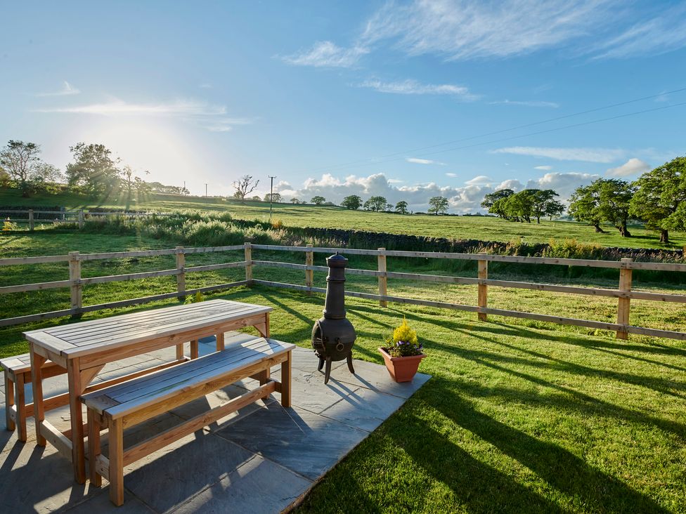 An outdoor seating area with a wooden table and benches a chiminea and a potted plant on a stone patio with a fenced grassy field and trees in the background at Whernside Lodge in Rathmell near Settle