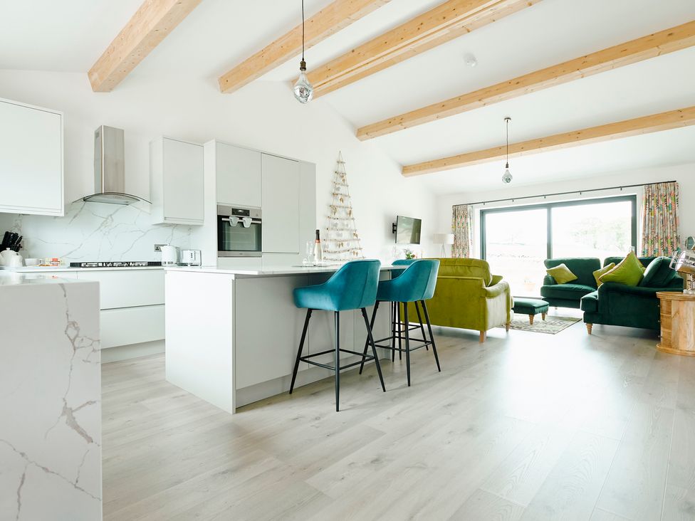 An open plan kitchen and living room with a white island and blue bar stools green sofas and wooden beams on the ceiling at Whernside Lodge in Rathmell near Settle
