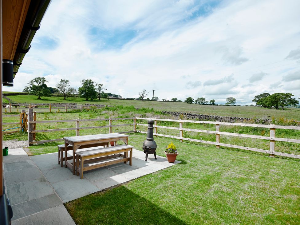 An outdoor patio with wooden picnic table and benches a chiminea and a potted plant on grass near a wooden fence with open field beyond at Whernside Lodge in Rathmell near Settle