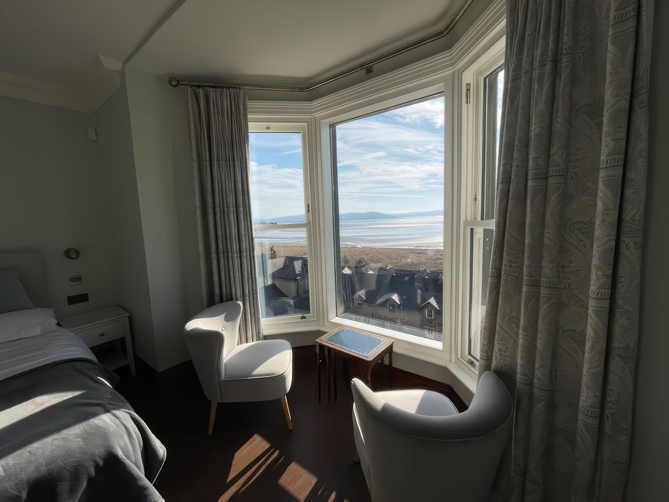 A bedroom corner with two chairs and a small table by a bay window overlooking rooftops and a beach at Victoria Mount in Grange-Over-Sands