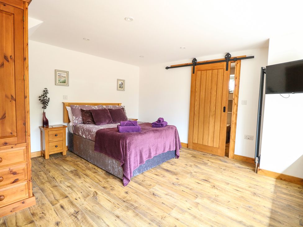 A bedroom with a double bed covered with purple bedding two wooden nightstands a wooden wardrobe a sliding wooden door and a wall-mounted television at Cobblers Barn in Repps With Bastwick near Potter Heigham